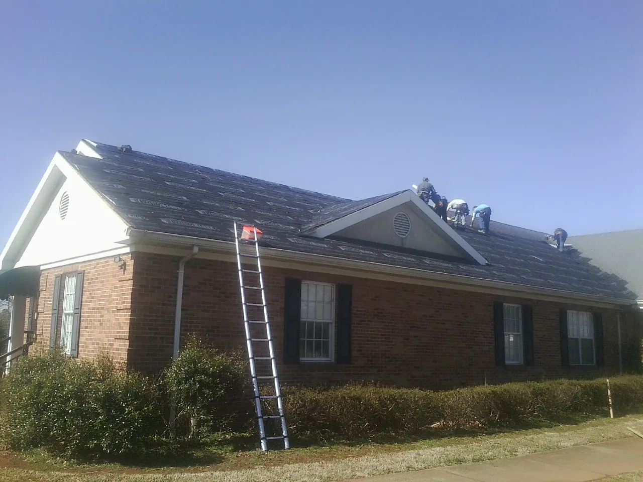 Crew tearing shingles off a brick ranch roof with a ladder set — Metal Roof Repair work by our crew in Staples